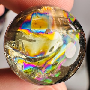 Può includere: Una sfera di cristallo trasparente e sferica con fratture interne e rifrazioni iridescenti arcobaleno. Il cristallo mostra una gamma di colori, tra cui giallo, blu e verde, creando un'esposizione vibrante. La sfera è tenuta in una mano.