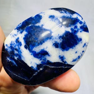 May include: A polished, oval-shaped sodalite stone featuring a marbled pattern of deep royal blue and opaque white sections. The surface is smooth and reflective, displaying natural mineral inclusions and a high-gloss finish throughout the piece.