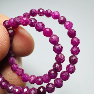 May include: A strand of small, faceted, deep magenta-colored gemstone beads arranged in a circular loop. Each bead features a multi-faceted surface that reflects light, creating a textured appearance across the vibrant, saturated pinkish-purple stones.