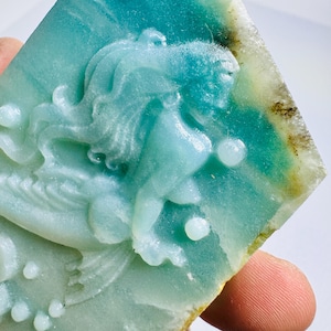 Puede incluir: Una talla rectangular de piedra turquesa que representa una sirena. La sirena tiene el pelo largo y suelto y está rodeada de burbujas. La piedra tiene un borde natural y rugoso con tonos verdes y marrones.