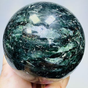 May include: A polished spherical stone featuring deep forest green and black mineral inclusions with a reflective, glossy surface. The orb displays intricate crystalline patterns and natural fissures throughout its dark, variegated texture.