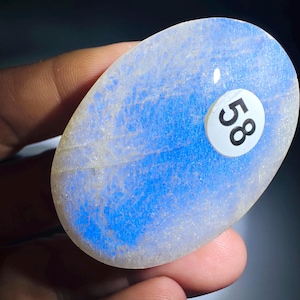 May include: An oval-shaped moonstone cabochon featuring a translucent white base with a vibrant blue adularescence effect. A small, circular white sticker with the black number 58 is adhered to the smooth, polished surface of the gemstone.