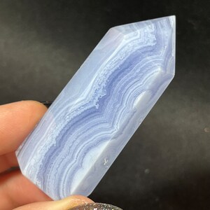 May include: A polished blue lace agate crystal tower featuring a hexagonal point and a smooth, translucent surface. The stone displays intricate, wavy white and light blue banding patterns throughout its length, creating a layered appearance.