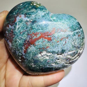 May include: A heart-shaped stone with a mottled pattern of green, red, and white. The stone has a smooth, polished surface and a rough, textured base. The colors are vibrant and the shape is symmetrical.