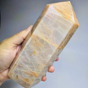 May include: A polished, hexagonal crystal tower featuring a translucent, pale beige and cream base color with prominent golden-brown, fibrous inclusions running throughout the stone. The crystal has a pointed, faceted top and a smooth, polished surface.