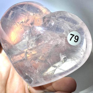 May include: A heart-shaped polished rose quartz crystal with a translucent, pale pink appearance. The stone features visible internal fractures and natural inclusions throughout its structure. A small white circular sticker with the number 79 is attached.