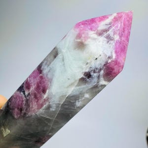 May include: A polished crystal point featuring a blend of vibrant magenta and deep pink inclusions contrasted against translucent white and grey mineral sections. The stone has a faceted, tapered top and a smooth, elongated, multi-toned body.