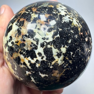 Puede incluir: Una piedra esférica pulida que presenta un patrón de alto contraste con inclusiones minerales negras angulares incrustadas en una matriz de color blanco crema y amarillo pálido, creando una textura compleja y moteada en toda la superficie.
