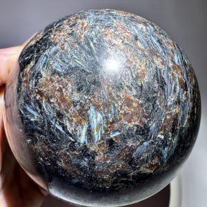 May include: A polished spherical stone featuring a complex texture of deep black, dark brown, and metallic blue mineral inclusions. The surface displays a reflective, crystalline pattern with streaks of iridescent blue light throughout.