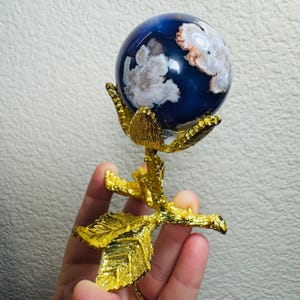 May include: A decorative sphere with a blue and white pattern, held by a gold-coloured metal rose. The sphere has a polished, glossy finish. The rose has detailed leaves and petals, creating an elegant display piece.