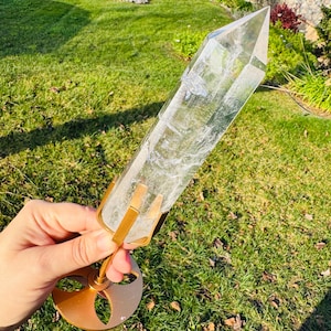 Può includere: Una grande punta di cristallo di quarzo trasparente montata su un supporto in metallo color oro. Il cristallo è tenuto in posizione da tre rebbi. Il supporto ha una base circolare decorativa con un design ritagliato. Il cristallo è alto circa 30 cm.