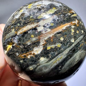 May include: A polished, spherical gemstone with a dark, mottled appearance. The stone features a mix of black, gray, brown, and yellow hues, with a banded pattern and small, yellow-green spots. The sphere is held in a hand.