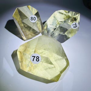 May include: Three faceted, translucent yellow crystals with geometric shapes. Each crystal features a white circular sticker with black printed numbers: 78, 79, and 80. The stones exhibit internal inclusions and sharp, polished edges that reflect light.