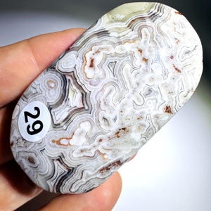 May include: A polished oval-shaped Crazy Lace Agate gemstone featuring intricate, swirling bands of white, cream, gray, and translucent tones with small reddish-brown inclusions. A circular white sticker with the black number 29 is attached.