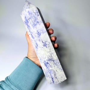 Op de afbeelding: Een hoge, rechthoekige kristallen obelisk met een puntige top. Het kristal heeft een gemarmerd patroon van witte en blauwe tinten. De obelisk wordt in een hand gehouden, wat de grootte en textuur laat zien. De kristal is ongeveer 20 cm hoog.