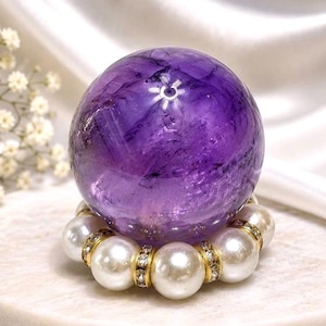 May include: A polished, deep purple amethyst sphere resting on a circular stand composed of white pearls and small, clear, sparkling rhinestones set in gold-toned metal, showcasing natural internal inclusions and a smooth, reflective surface.