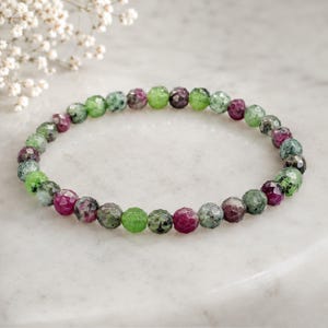 Könnte beinhalten: Ein Perlenarmband aus kleinen, runden, facettierten Steinen in abwechselnden Farbtönen von tiefem Magenta, Waldgrün und gesprenkeltem Graugrün. Die Steine sind in einem durchgehenden kreisförmigen Muster aufgereiht und bilden eine texturierte Oberfläche.