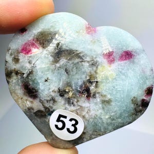 May include: A heart-shaped polished stone featuring a pale blue and translucent base with embedded dark grey mineral inclusions and vibrant magenta-pink crystal patches. A small white circular sticker with the black number 53 is attached.