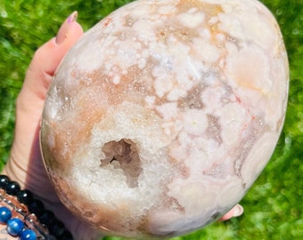 Puede incluir: Una gran geoda de ágata rosa y blanca con una abertura natural que revela un interior forrado de cristales. La geoda tiene un exterior liso y pulido y un patrón único y arremolinado.