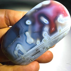 May include: A polished, rounded agate stone featuring a smooth surface with translucent layers of deep blue, purple, and red. The stone displays distinct white circular inclusions and linear banding patterns throughout its surface.