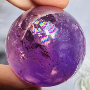 May include: A polished, spherical amethyst crystal ball. The translucent purple sphere has internal inclusions and a rainbow-colored flash of light refracting within. The crystal is held between two fingers, showcasing its smooth, rounded form.