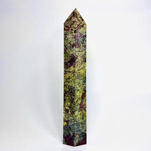May include: A tall, rectangular stone obelisk with a pointed top. The obelisk features a mottled pattern of green, burgundy, and yellow hues. The stone has a textured appearance, and the obelisk stands upright against a white background.