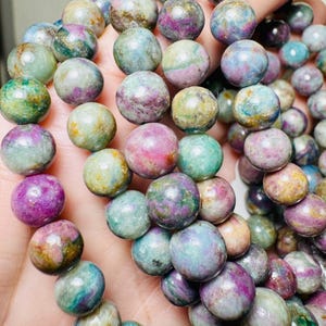 Könnte beinhalten: Ein Armband mit runden, polierten Edelsteinen in Lila, Grün, Blau und Rosa. Die Perlen haben ein marmoriertes Aussehen mit Farb- und Texturvariationen. Das Armband ist aufgefädelt und zeigt die natürliche Schönheit der Steine.