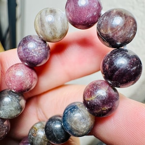 May include: A beaded bracelet featuring a variety of polished, round gemstones in shades of purple, gray, and blue. The beads are strung together, creating a bracelet. The bracelet is held by a person's fingers.