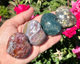 May include: Four polished heart-shaped stones arranged in a row. The first is a mottled reddish-brown, the second is a translucent lavender and grey, the third is a deep forest green with dark flecks, and the fourth is a dark brown with cream spots.
