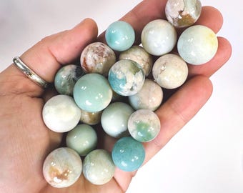 May include: A collection of polished gemstone spheres in various colors, including light blue, cream, and brown. The spheres are held in a hand, showcasing their smooth surfaces and natural patterns. The gemstones are approximately 1 inch in diameter.