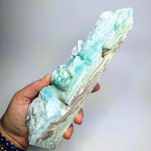 May include: A large, elongated raw mineral specimen featuring vibrant turquoise and light blue crystalline formations with layered, textured white and tan base rock. The stone displays a natural, jagged surface with varied mineral deposits.