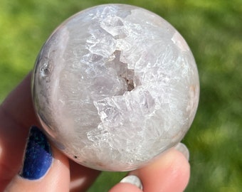 May include: A white agate sphere with a large, open geode inside. The geode is filled with smaller, white crystals. The sphere is smooth and polished.