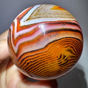 May include: A polished, spherical agate stone with a vibrant display of colors. The orb features bands of orange, white, pink, and black, creating a layered, swirling pattern. The stone's surface is smooth and reflective, showcasing its natural beauty.