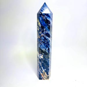 May include: A tall, six-sided crystal tower featuring a polished finish. The stone displays a marbled pattern of deep royal blue, navy, and white, with subtle streaks of dark grey and brown mineral inclusions throughout its vertical structure.