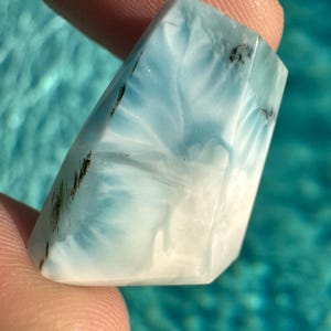 May include: A polished, faceted gemstone with a light blue and white color gradient. The stone has dark inclusions and a unique pattern resembling ocean waves. The gemstone is held between two fingers, showcasing its shape and color.