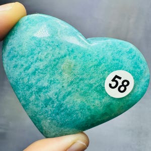 May include: A heart-shaped, polished stone in shades of teal and green. The stone has a smooth, glossy surface with natural variations in color. A white sticker with the number "58" is affixed to the stone.