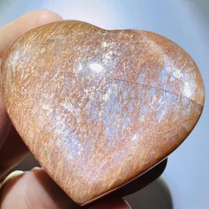 May include: A polished heart-shaped sunstone featuring a warm, golden-brown hue with shimmering, reflective inclusions throughout the surface. The stone displays a smooth, rounded texture with visible internal mineral layers.