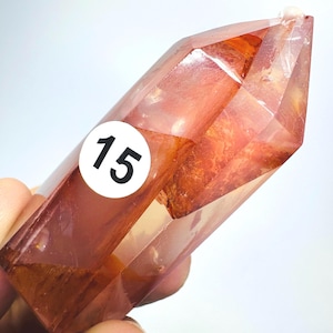 May include: A polished, faceted crystal point with a translucent, deep orange and reddish-brown hue. The crystal features internal inclusions and a distinct, sharp termination at the top. A circular white sticker with the black number 15 is attached.