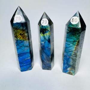 May include: Three polished labradorite crystal towers standing upright. Each stone features a dark gray base with iridescent blue and yellow flashes. White circular stickers labeled 78, 79, and 80 are affixed to the upper front of each crystal point.