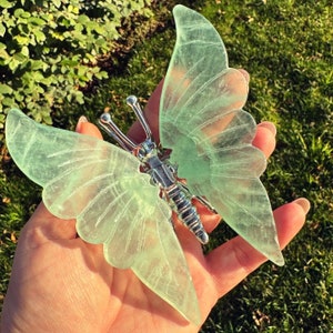 Puede incluir: Una escultura de mariposa translúcida verde claro con patrones de alas detallados y un cuerpo plateado. La mariposa se sostiene en una mano sobre un fondo de hierba verde y follaje, mostrando su delicado diseño.