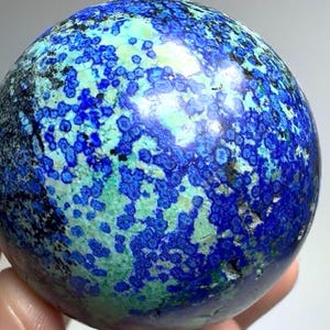 May include: A polished spherical stone featuring a vibrant mix of deep royal blue and bright turquoise green mineral patterns. The surface displays a mottled, speckled texture with small dark black inclusions scattered throughout the rounded form.