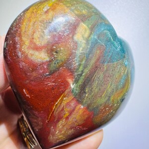 May include: A polished heart-shaped stone featuring a vibrant mix of colors including deep red, golden yellow, forest green, and bright blue. The surface is smooth and glossy, displaying natural swirling patterns and mineral inclusions throughout.