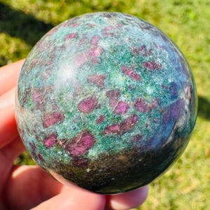 Peut inclure: Une sphère polie de rubis zoïsite, présentant un mélange de teintes vert foncé, sarcelle et violet. La surface est parsemée de taches rouge rubis vives et irrégulières, créant un contraste saisissant. La pierre est tenue dans une main.