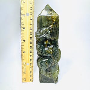 May include: A dark green labradorite crystal tower carved with a dragon figure, positioned next to a yellow ruler showing a height of approximately 7 inches. The stone features natural inclusions and a polished finish with visible dragon scales.