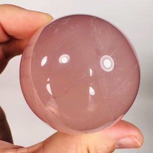 May include: A translucent, rose-colored crystal sphere. The polished surface reflects light, creating bright highlights. The sphere is held in a hand, showcasing its smooth, rounded form. This item is a decorative crystal ball.