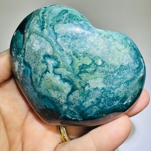 May include: A heart-shaped stone with a swirling pattern of teal, green, and blue hues. The polished surface has a glossy sheen, with hints of red and white. The stone is held in a hand, showcasing its natural beauty.