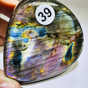 May include: A polished labradorite stone with a smooth, rounded triangular shape. The surface displays iridescent flashes of blue, purple, and yellow against a dark gray base with visible internal fractures. A white circular sticker with the number 39 is attached to the top.