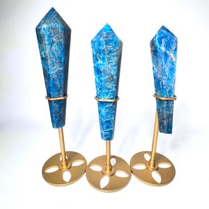 May include: Three blue apatite crystal points on gold-colored metal stands. The crystals have a faceted, elongated shape with a pointed top and are secured by a gold ring. The stands have a circular base with a floral design.