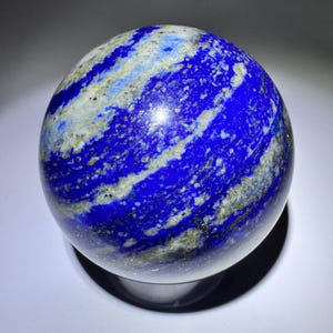 May include: A polished lapis lazuli sphere, displaying rich blue tones with white and gold veining. The gemstone ball sits on a dark stand, reflecting light. A decorative and unique item, approximately 8 cm in diameter.