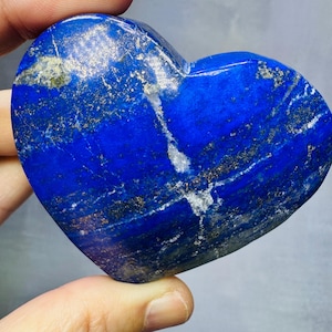 Op de afbeelding: Een hartvormige lapis lazuli steen. De steen heeft een diepblauwe kleur met witte strepen en gouden vlekken. Het hart is glad en gepolijst en wordt in de hand van een persoon gehouden.